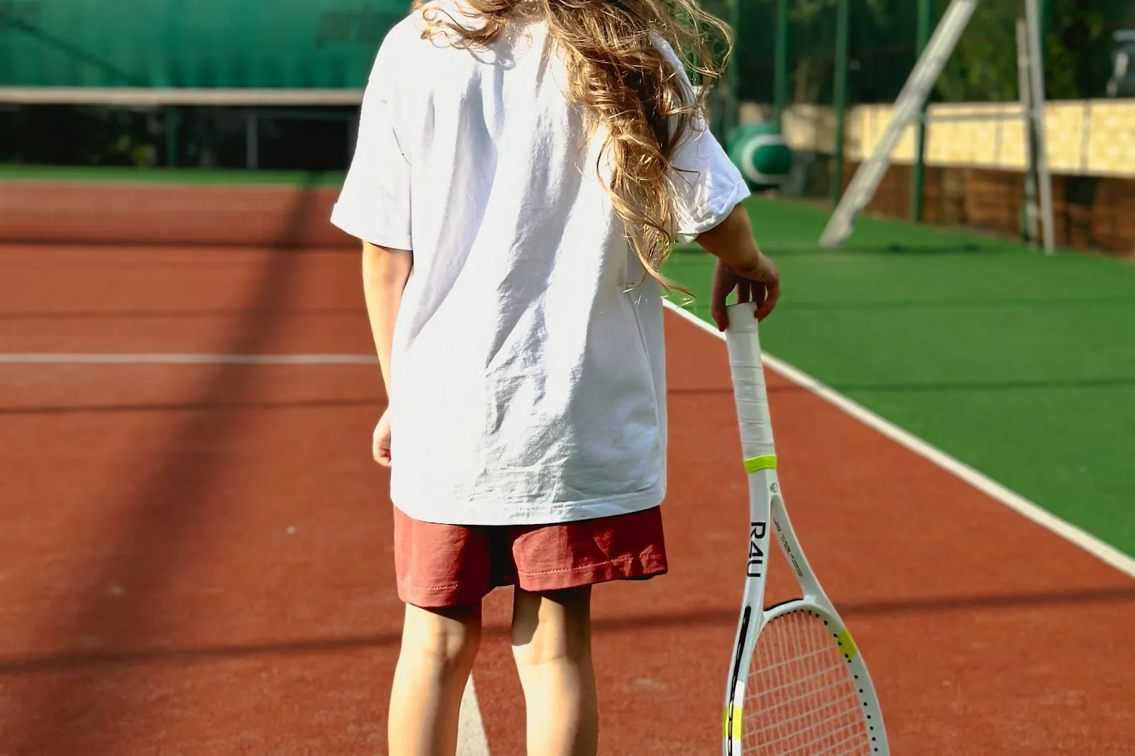 Une enfant vue de dos, appuyée sur sa raquette, seule sur un court de tennis en terre battue.