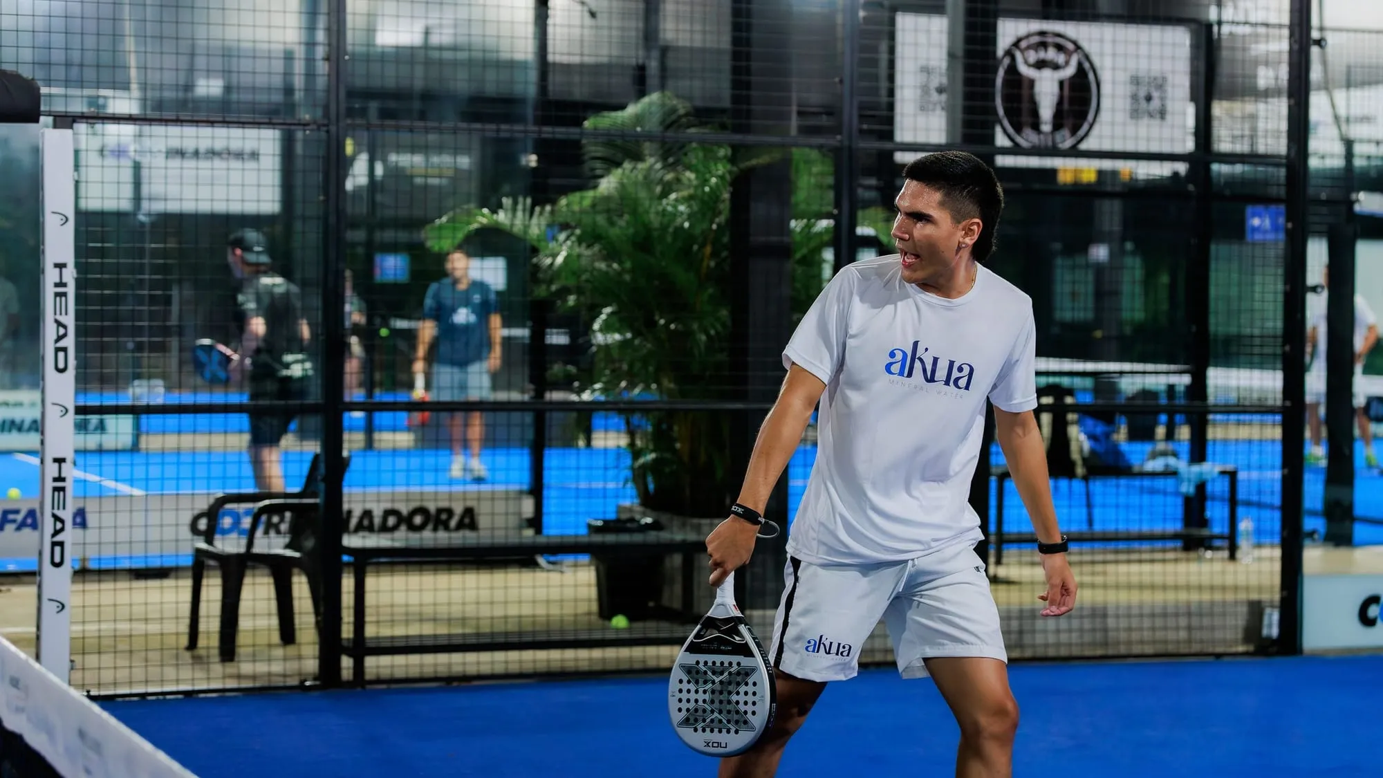 Un joueur de padel raquette en main pendant une partie en nocturne, sur un terrain vitré éclairé.