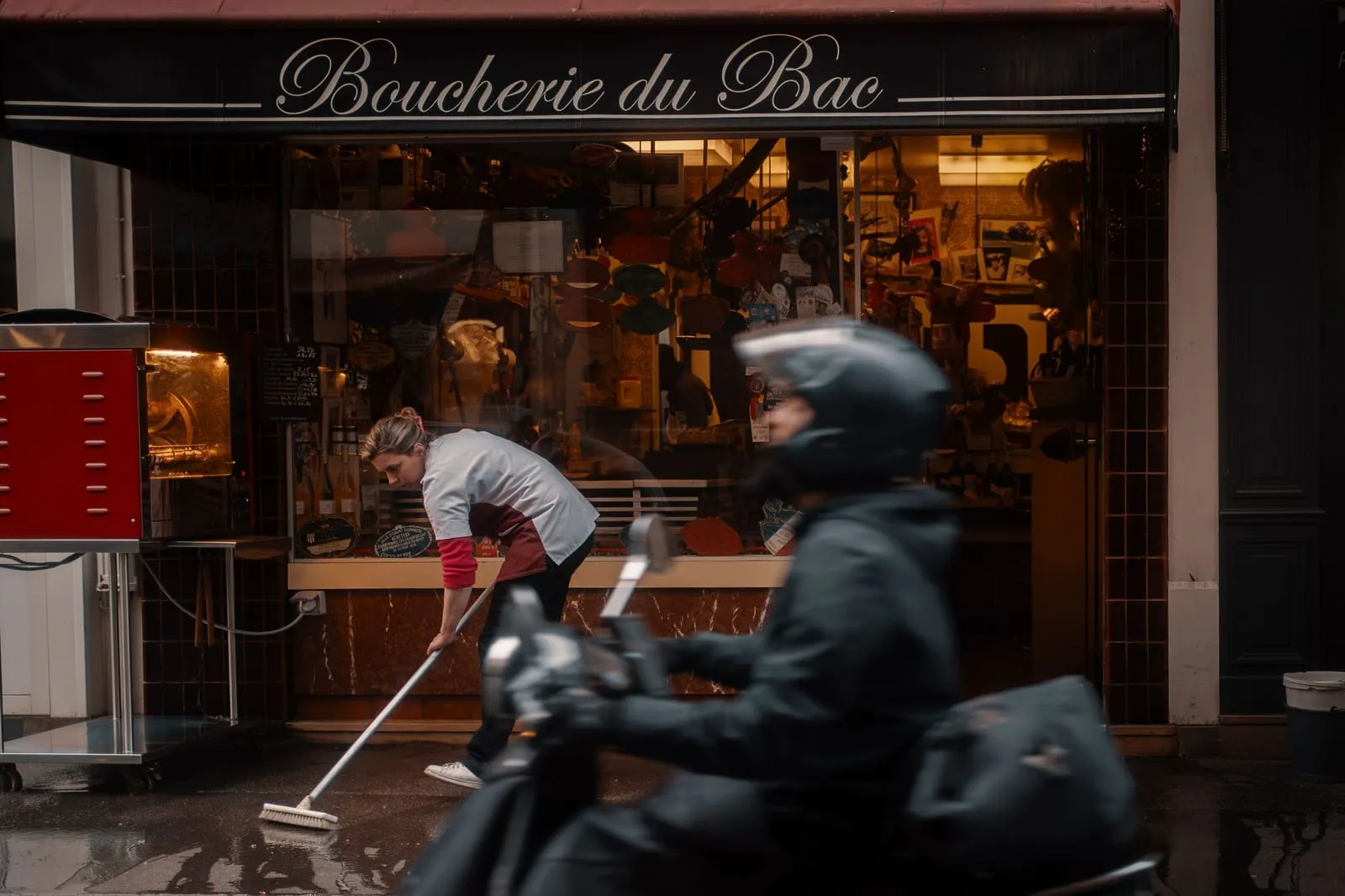 La devanture éclairée d'une boucherie de quartier au petit matin, un employé balaie devant la vitrine.